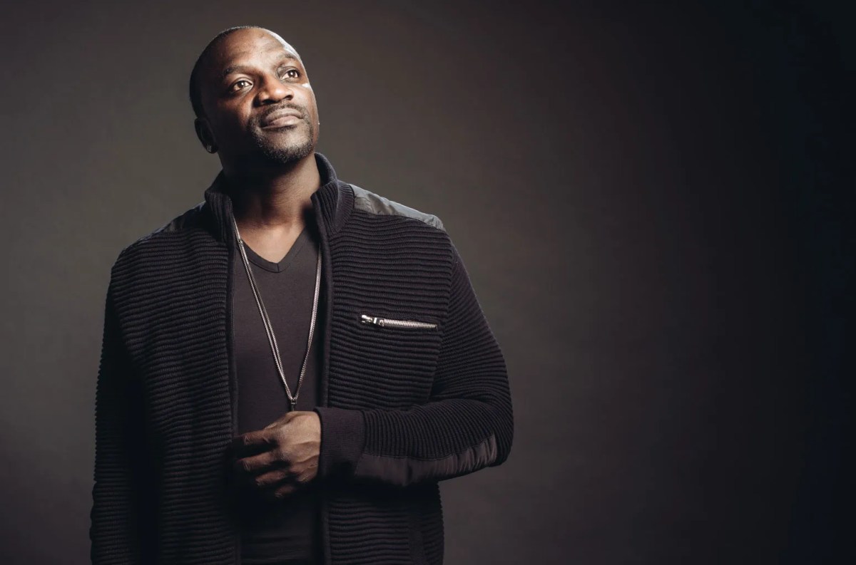 Akon stands in front of a black background in a black suit.