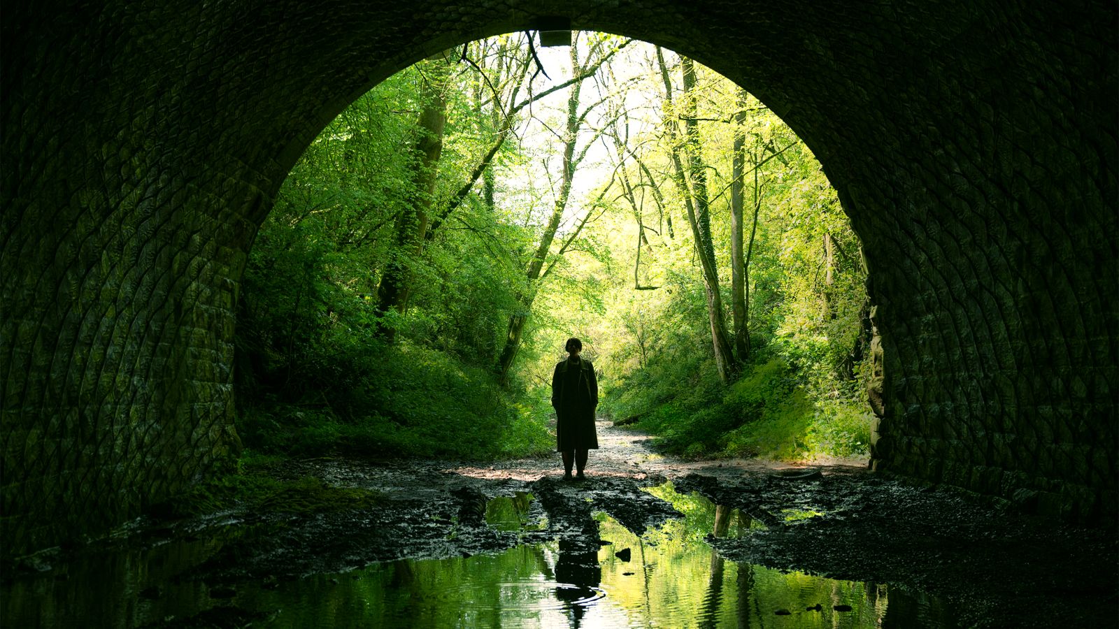 In a still from ‘Men,’ a dark figure of a person in the distance silhouetted against a spooky/beautiful green forest landscape