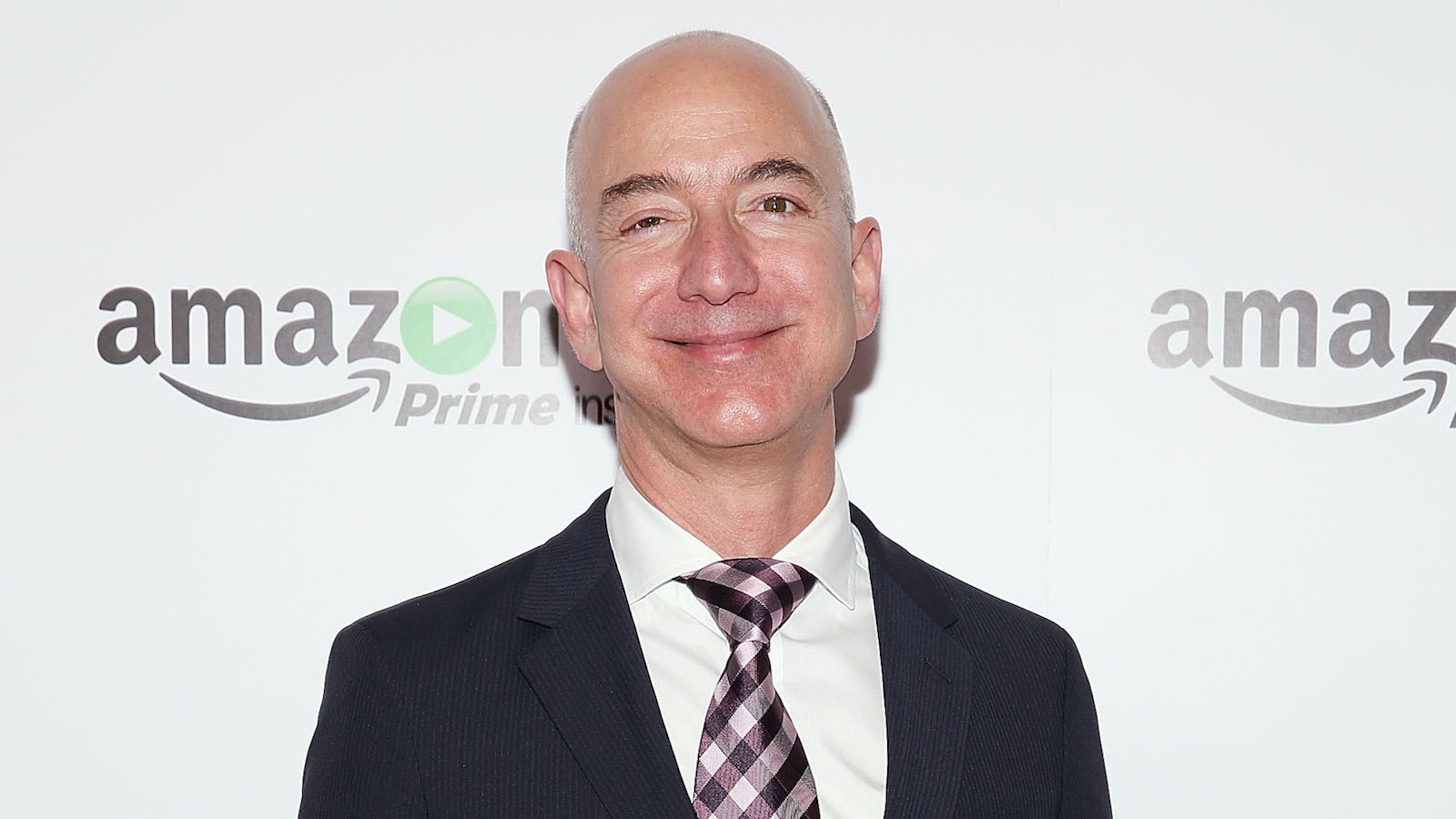 Jeff Bezos in a black and white suite, purple checkered tie, and smiling with his mouth closed. 