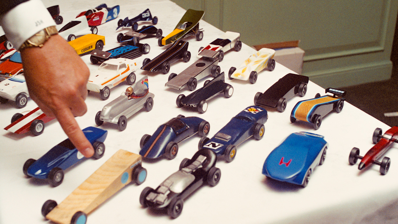 A man’s hand points at one of numerous vintage toy cars on a white tablecloth.