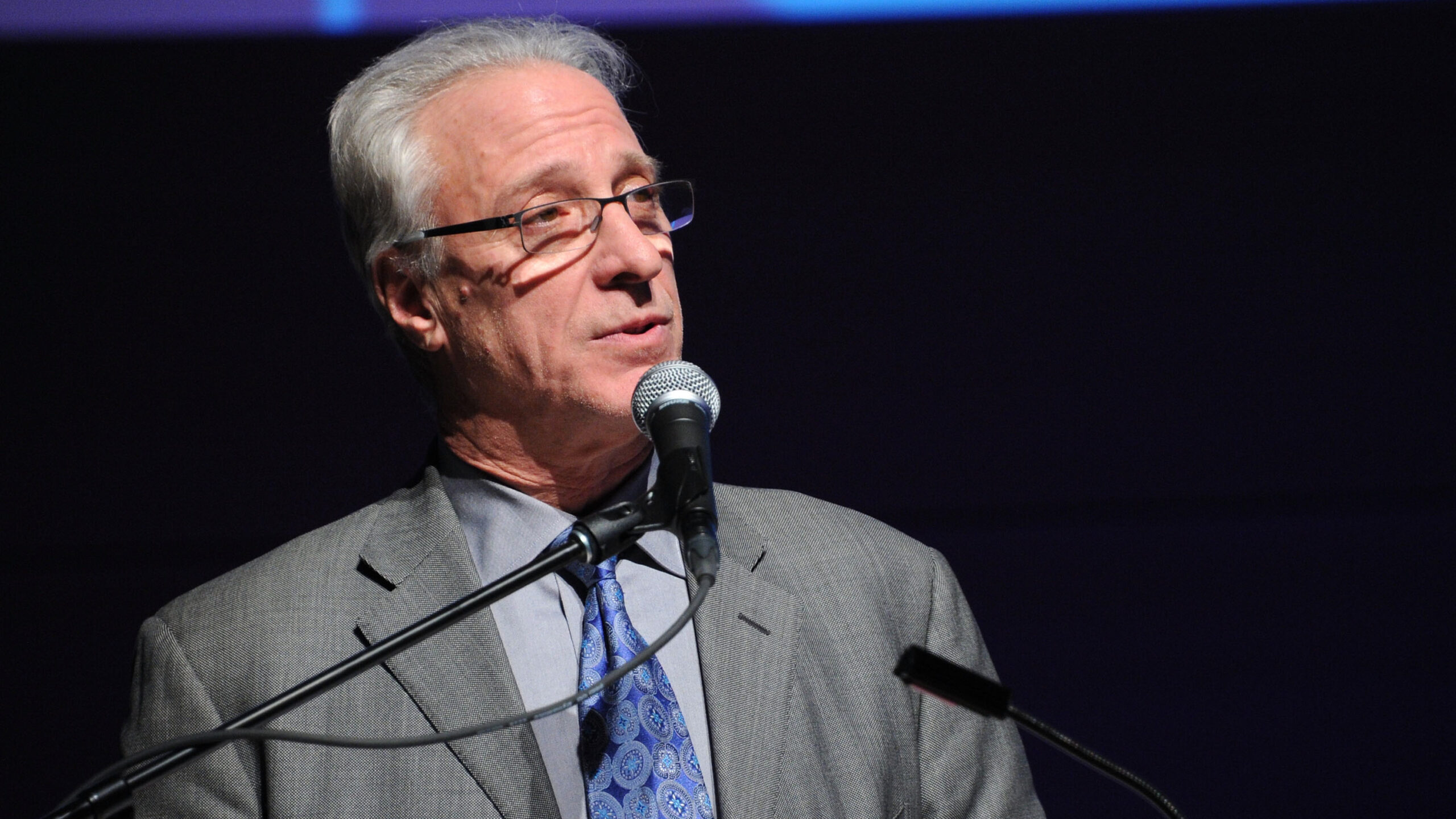 The actor gives a speech, standing at a podium/microphone, looking thoughtfully off-camera