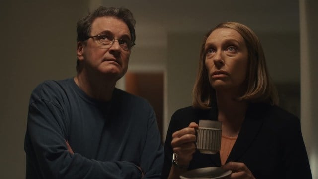 Colin Firth and Toni Collette stand together gazing upward in a dimly lit hall