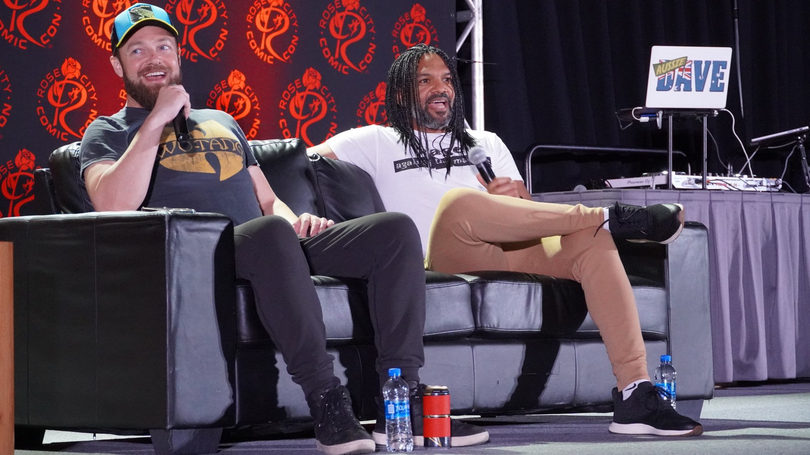 Khary Payton and Ross Marquand field fan questions from a couch on a stage at a Comic Con panel in casual dress.