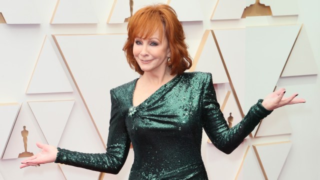 Reba McEntire makes a shrug pose in a green sequin dress at a red carpet event.
