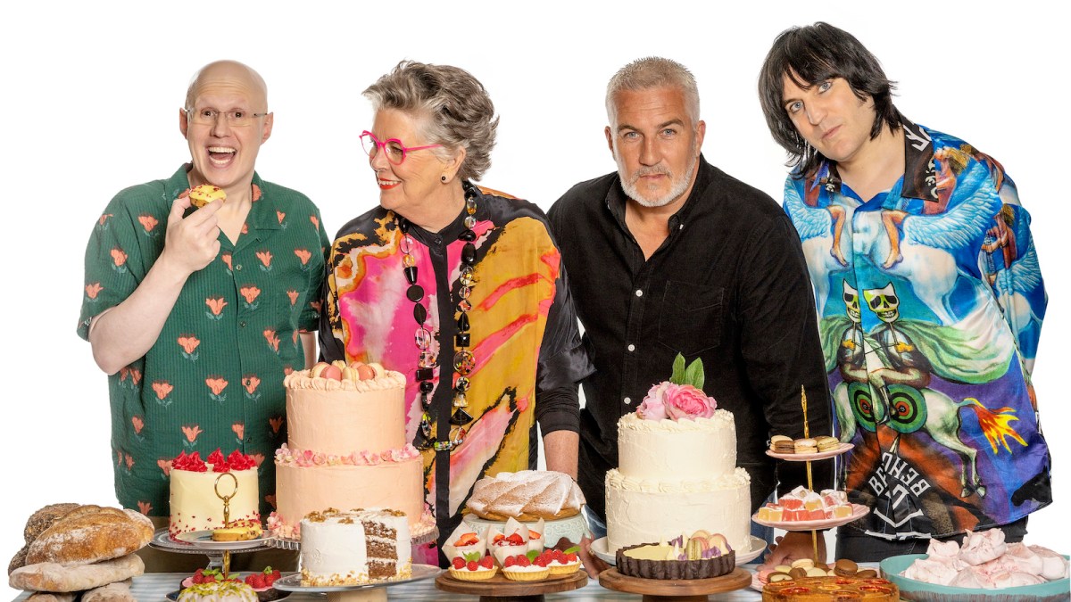 The hosts of The Great British Baking Show stand behind a table full of baked goods