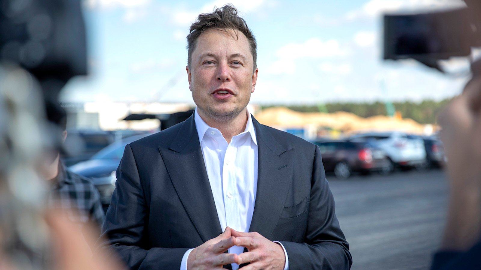 Elon Musk in a black and white suit speaking to a barrage of camera pointed at him