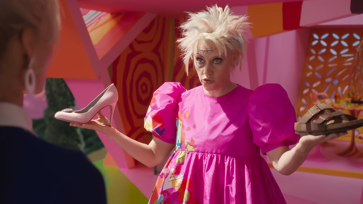 Kate McKinnon as Weird Barbie is holding up two different shoes.