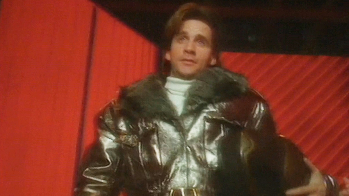 Ace Rimmer strolls triumphantly down a hallway wearing a bomber jacket and clutching his pilot's helmet. His long flowing hair is accompanied by a reassured grin.