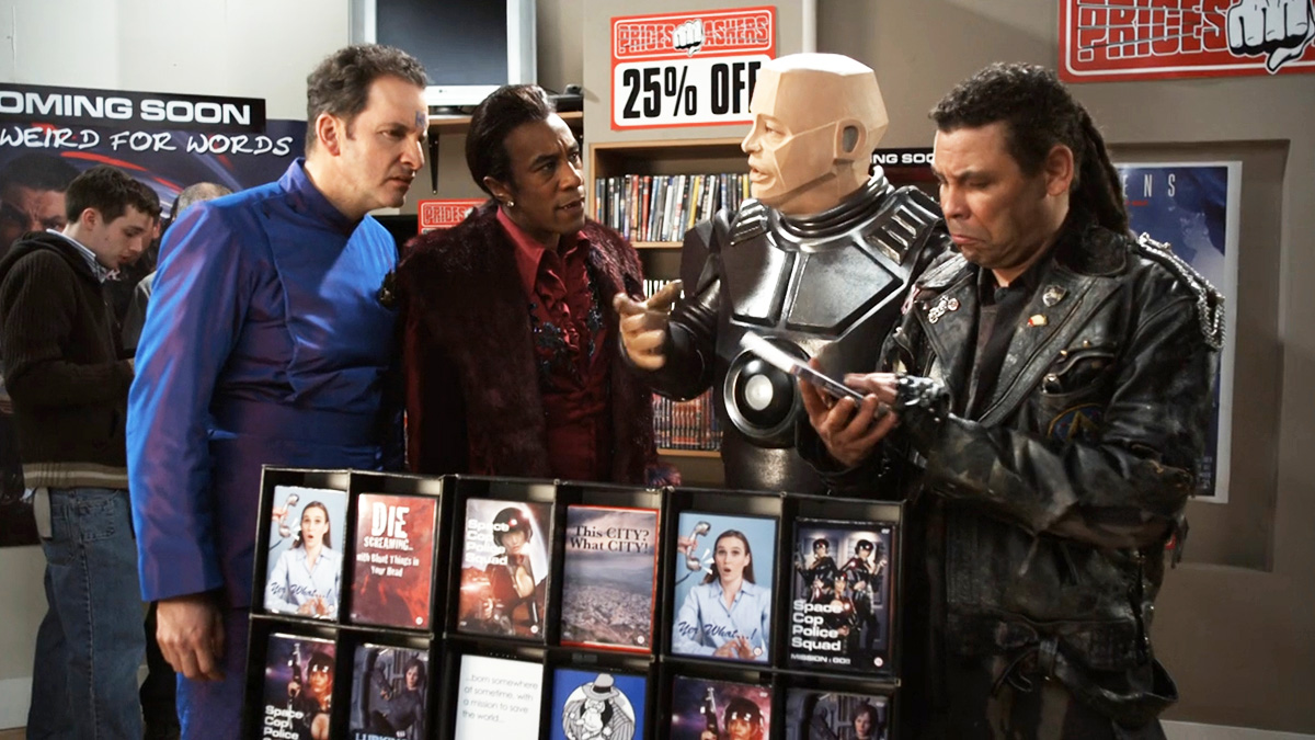 Rimmer, the Cat, Kryten, and Lister give puzzled looks at a DVD collection of their own adventures inside a video store.