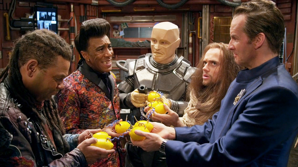 Lister, the Cat, Kryten, Jesus, and Rimmer stand in a circle as they all clutch lemons held together by circuitry. 