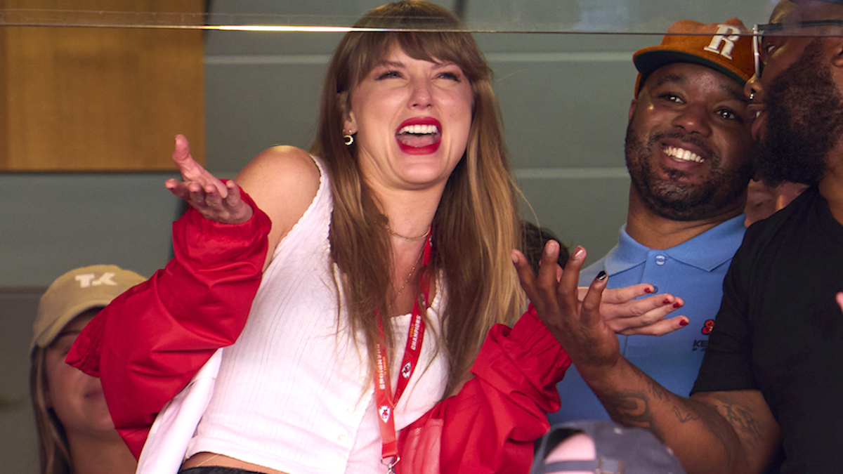 Taylor Swift shrugging and laughing while wearing a white shirt and red jacket in the box seats at the Kansas City Chiefs game