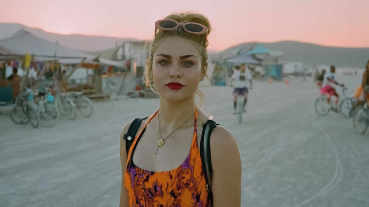 A blond woman with her hair put up stands on a beach, looking towards the camera.