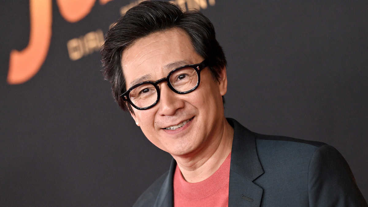 Ke Huy Quan in a black blazer, orange shirt, and standing in front of a black backdrop on the red carpet