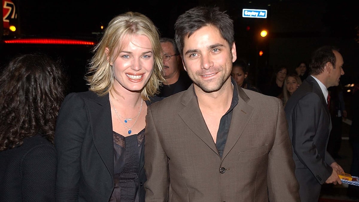 Rebecca Romijn and John Stamos pose for a photo together, smiling. 