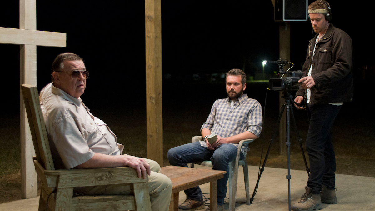 Man sits in front of a cross, being interviewed by two men across from him. 