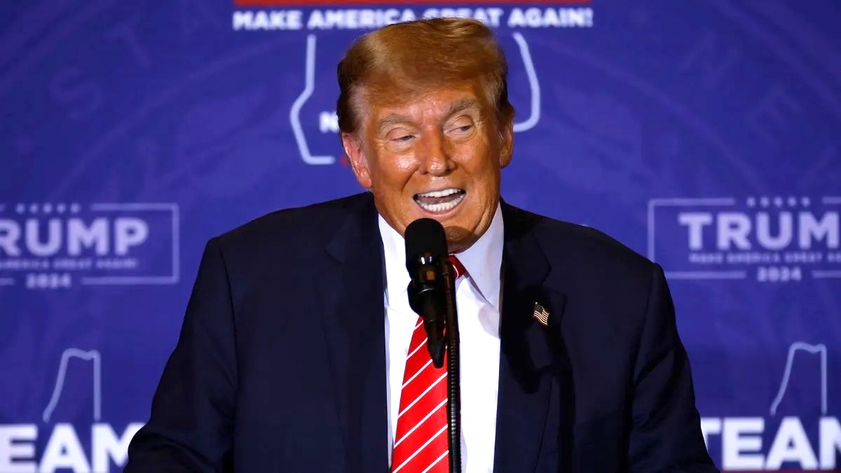 Donald Trump wearing a dark suit jacket, red tie, and standing at a microphone in front of a blue background with the Trump logo