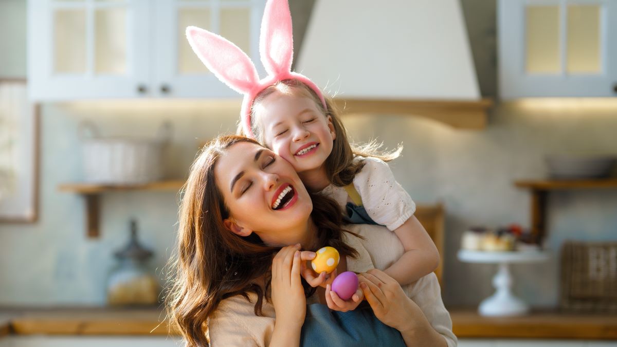 Happy mother and her daughter wearing aprons holding painted colorful eggs while decorating them with food dyes in cozy kitchen at home. Easter craft activities for families.