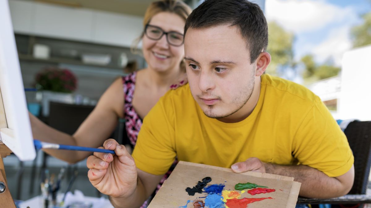 Teen boy with Down syndrome painting on canvas with his tutor on terrace above riverbank