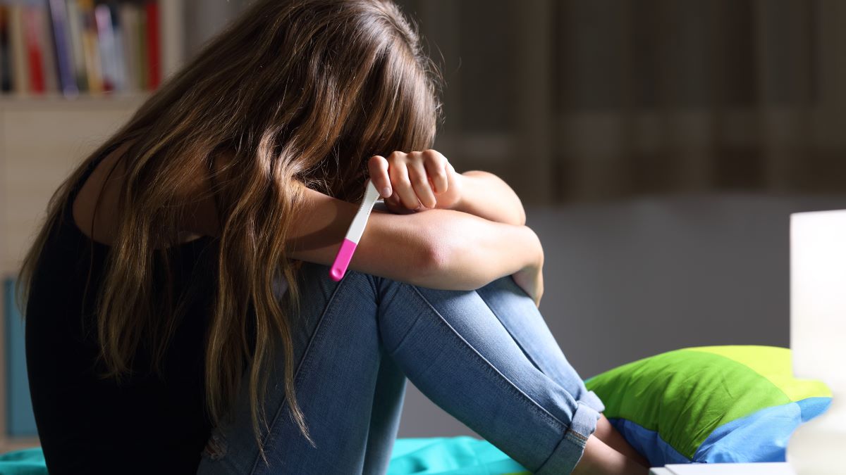 Side view of a sad pregnant teen sitting on her bed after checking a pregnancy test with a dark light in the background