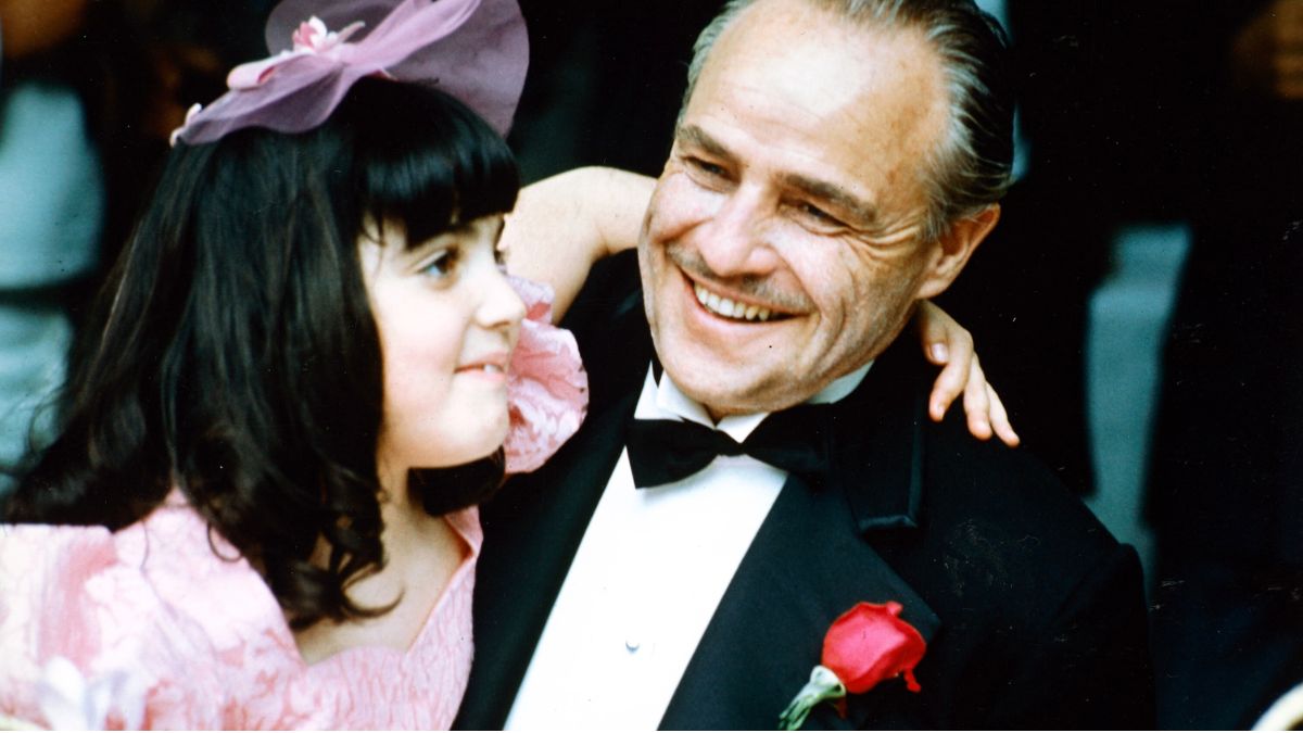 Marlon Brando (19242004), US actor, smiling and wearing a black dinner suit with a white shirt, with wing-tip collars, and a black bow tie, with a young girl in a pink dress, who has her arm around him, in a publicity still issued for the film, 'The Godfather', 1972. The mafia drama, directed by Francis Ford Coppola, starred Brando as 'Don Vito Corleone'. (Photo by Silver Screen Collection/Getty Images)